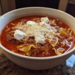 A close-up of a white bowl filled with rich, red Lasagna Soup, topped with ground meat, melted mozzarella, and dollops of ricotta cheese.