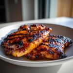 Close-up of three pieces of perfectly Grilled Hot Honey Chicken with a dark, sticky glaze and char marks, served on a white plate.