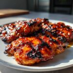 Close-up of three pieces of perfectly caramelized Grilled Hot Honey Chicken with dark char marks on a light gray plate.