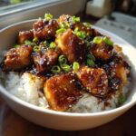 Close-up of a white bowl filled with white rice topped with glazed, saucy Honey BBQ Chicken Rice Bowl pieces, garnished with sesame seeds and green onions.