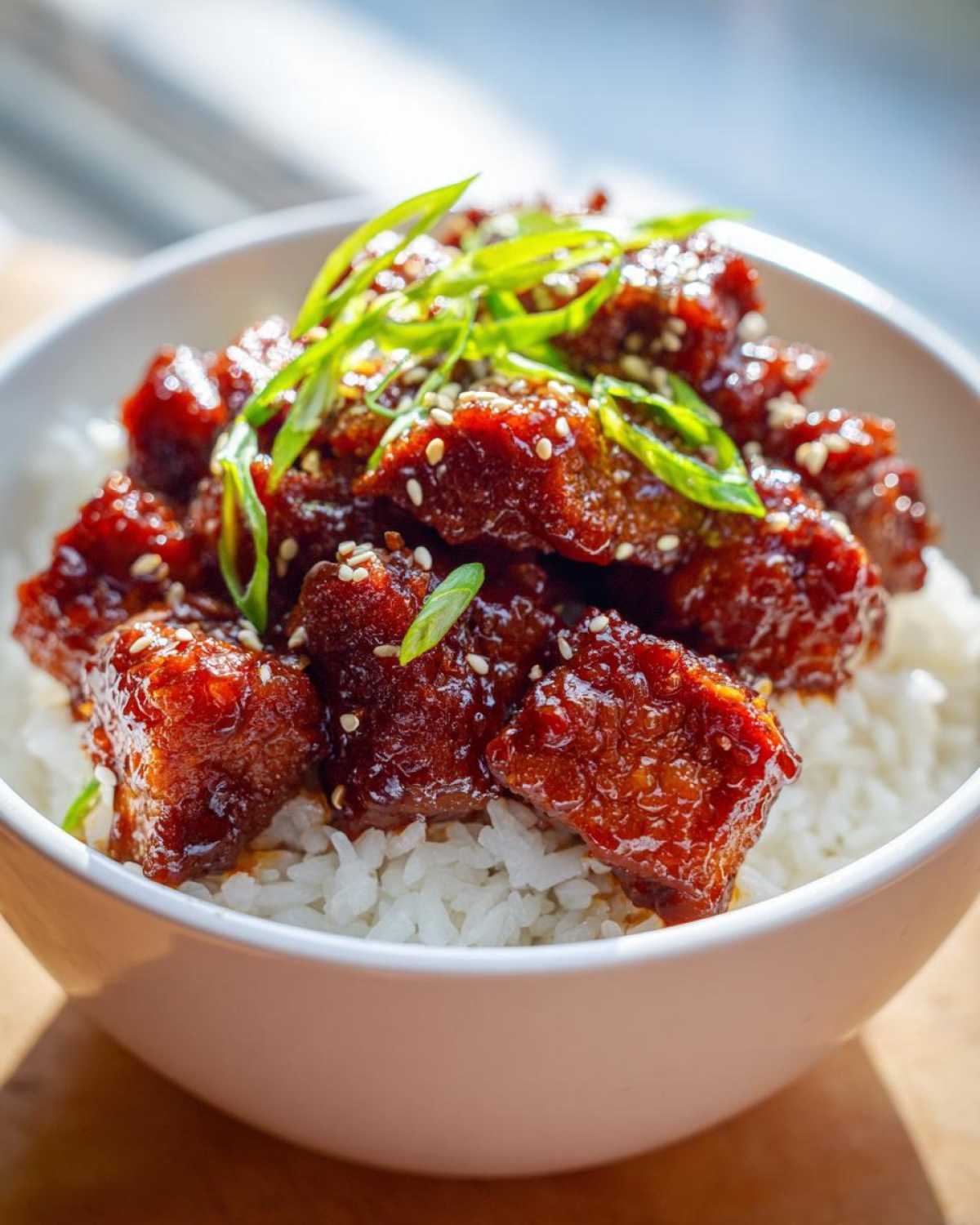 Close-up of a Honey BBQ Chicken Rice Bowl featuring sticky, dark red glazed chicken pieces over white rice, garnished with sesame seeds and green onions.