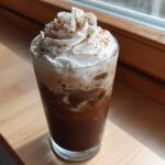 A tall glass of Iced Banana Bread Latte topped with whipped cream and cinnamon.