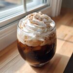 A tall glass of Iced Banana Bread Latte topped with whipped cream and cinnamon.