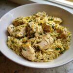 A close-up of a white bowl filled with Lemon Chicken Orzo Stir Fry, garnished with fresh parsley and lemon zest.