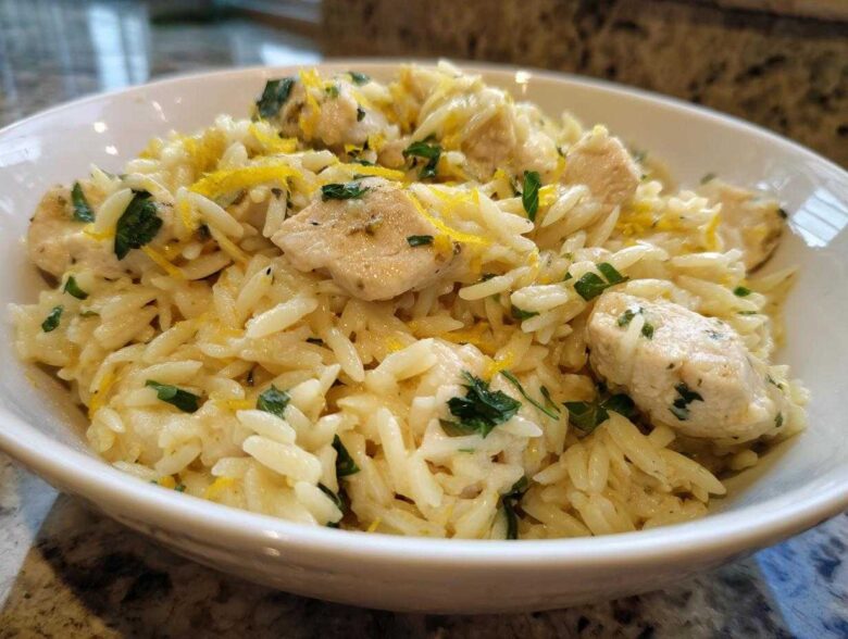 Close-up of a white bowl filled with Lemon Chicken Orzo Stir Fry, garnished with lemon zest and parsley.
