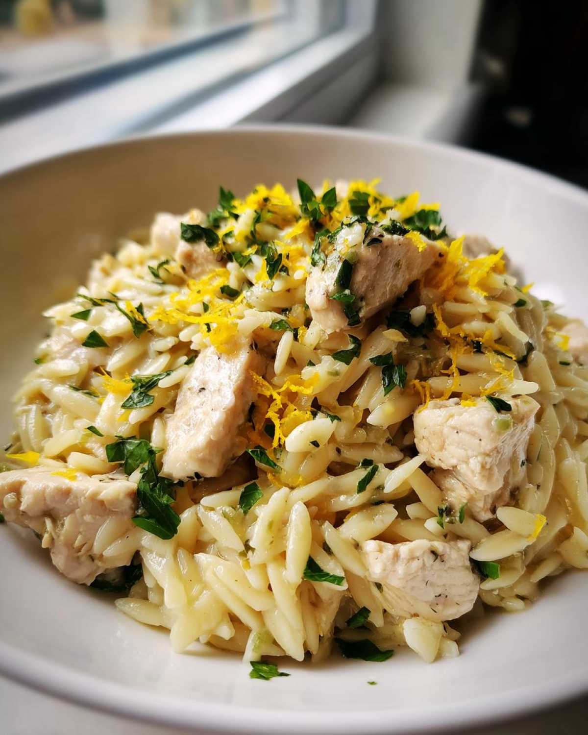A close-up of a white bowl filled with Lemon Chicken Orzo Stir Fry, topped with lemon zest and fresh parsley.