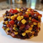 A perfectly molded serving of Dense Bean Salad featuring black beans, yellow corn, red peppers, and red onion on a white plate.