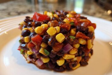 A perfectly molded serving of Dense Bean Salad featuring black beans, yellow corn, red peppers, and red onion on a white plate.
