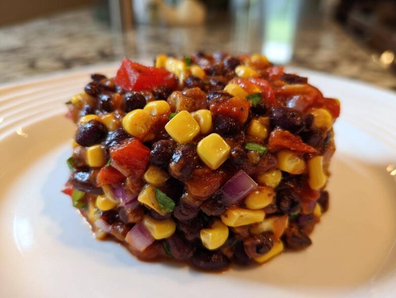 A perfectly molded serving of Dense Bean Salad featuring black beans, yellow corn, red peppers, and red onion on a white plate.