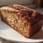 A close-up of a thick slice of Snickerdoodle Banana Bread with a dark, caramelized crust and a visible banana slice on top.