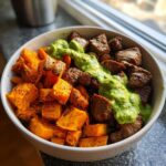 A close-up of a bowl featuring seasoned steak cubes, roasted sweet potatoes, and a vibrant avocado-cilantro drizzle.