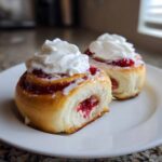 Close-up of two Strawberry Shortcake Cheesecake Rolls topped with bright red filling and a swirl of whipped cream on a white plate.